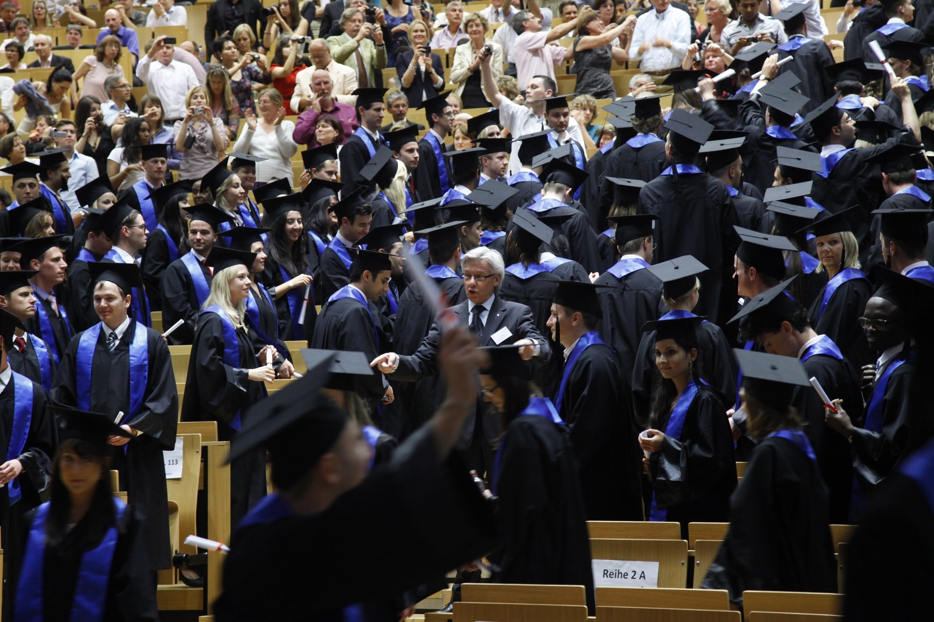Graduates in black gowns with blue stoles in a lecture hall, holding their certificates.