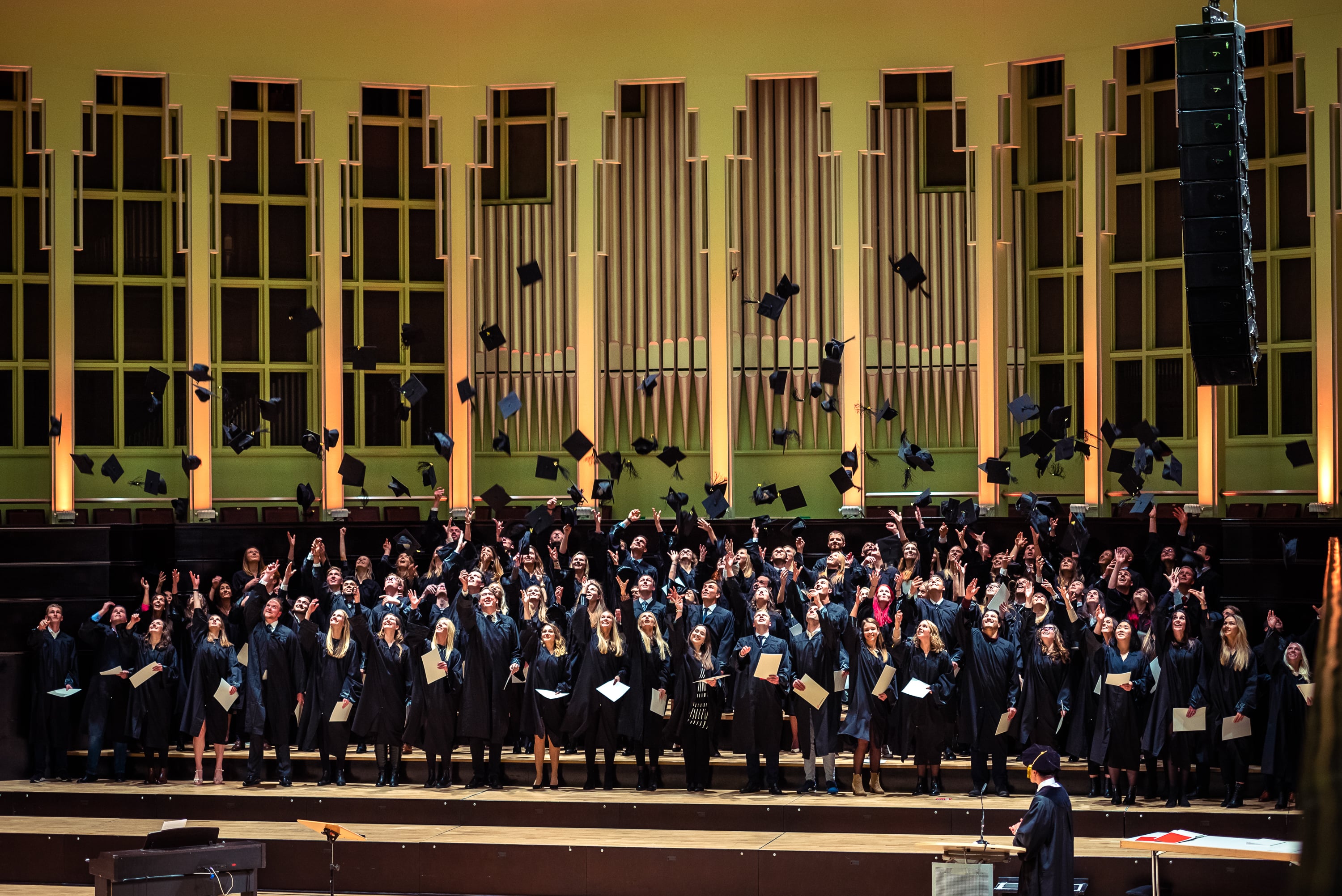 Graduation ceremony in a concert hall: a whole cohort in black gowns throws their caps into the air, degree certificates in hand.